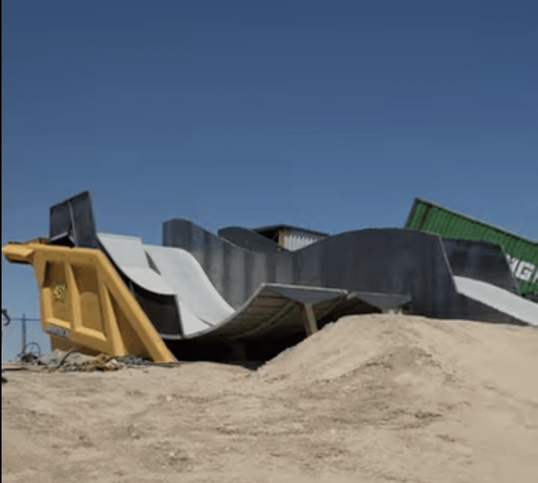 A modern concrete skate ramp with curved and angular sections is built on a dirt lot under a clear blue sky. A green shipping container is partially visible in the background.